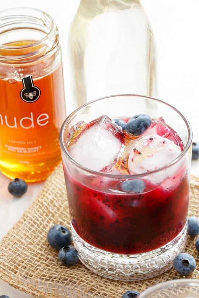 Blueberry-Honey Whiskey Sour in glass cup with ice, honey, glass bottle and blueberries around