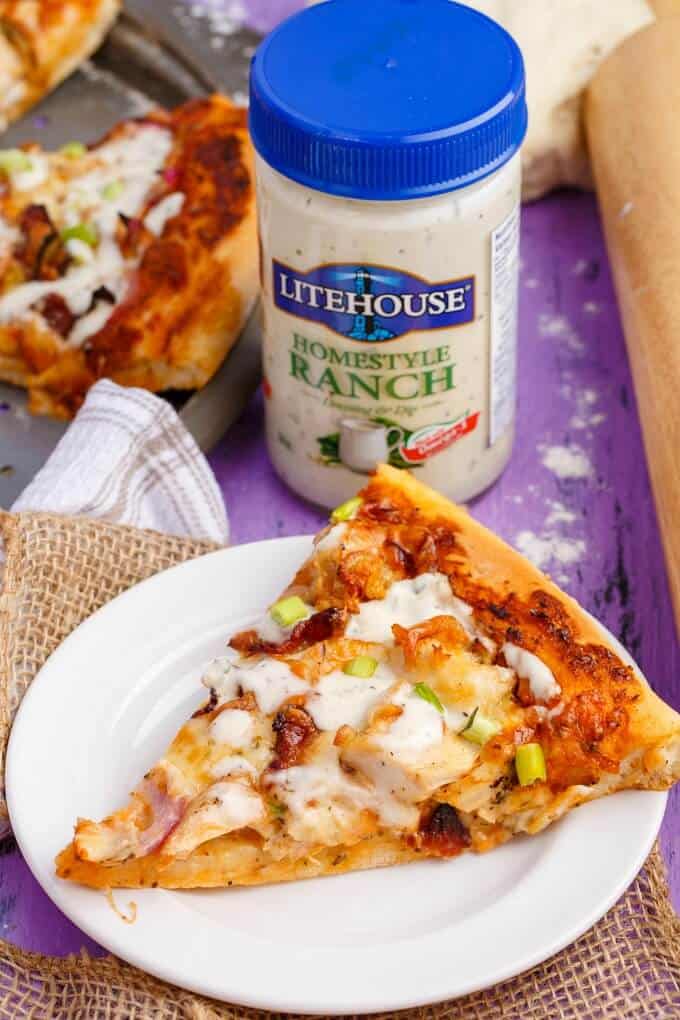 Chicken Bacon Ranch Pizza on white plate and gray tray, Litehouse dressing in jar with woden roller and pizza dough on purple table