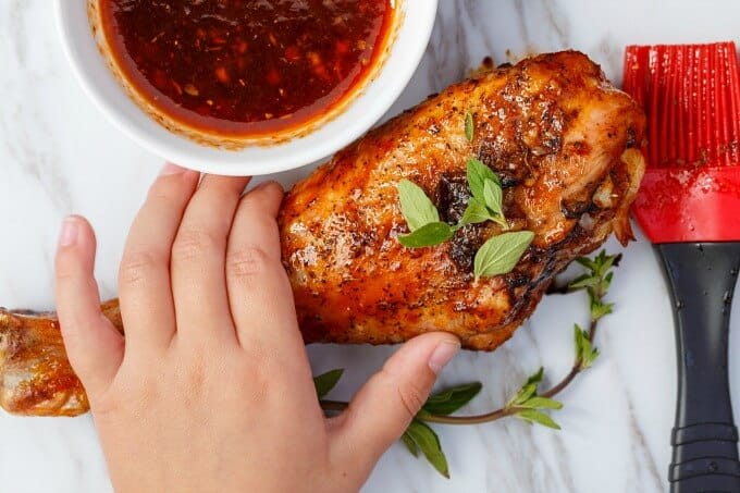 Grilled Sriracha Turkey Drumsticks held by hand, sauce in white bowl with red brush on the table
