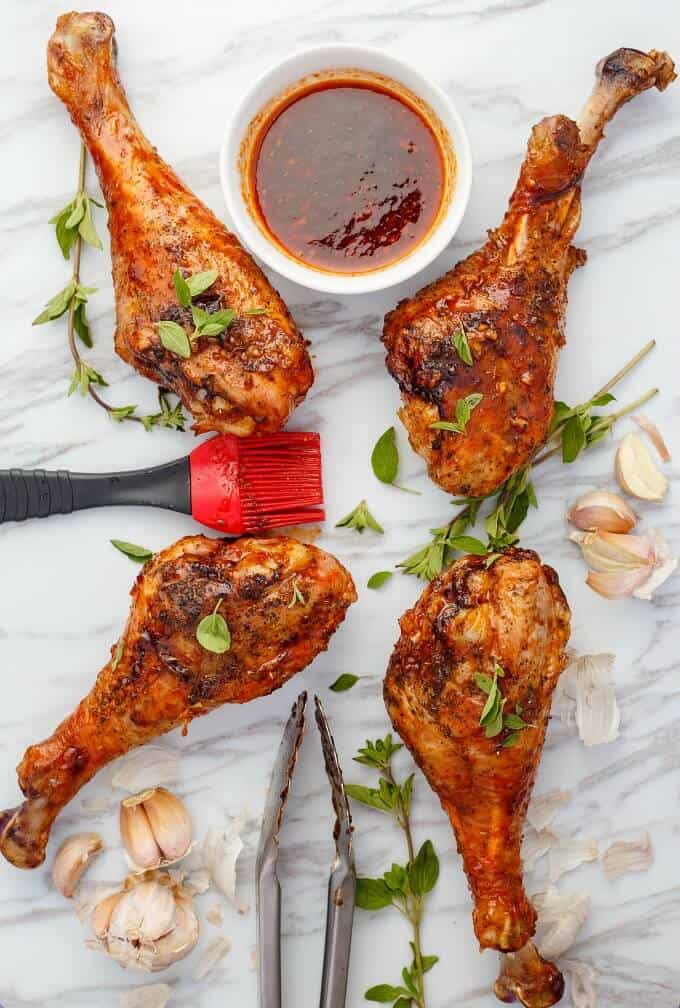 Grilled Sriracha Turkey Drumsticks with red brush, herbs, garlic, bowl of sauce and kitchen tongs