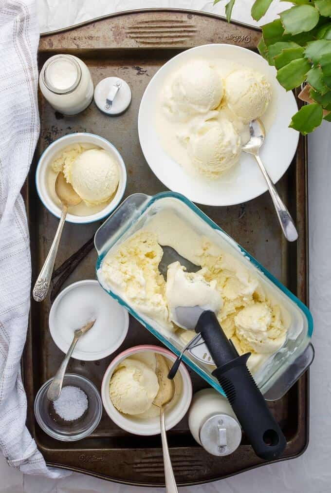 Homemade Vanilla Bean Ice Cream in glass container, bowls and white plate with spoons, ingredients in small bowls, jars of milk on gray tray