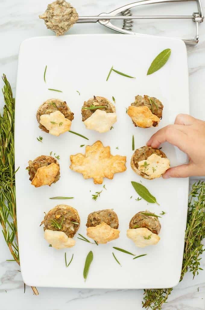 Tourtiere Stuffed Mushrooms  on white tray with herbs, with kitchen tool on table. Hand touching one piece of pie (Meat Pie Mushrooms)