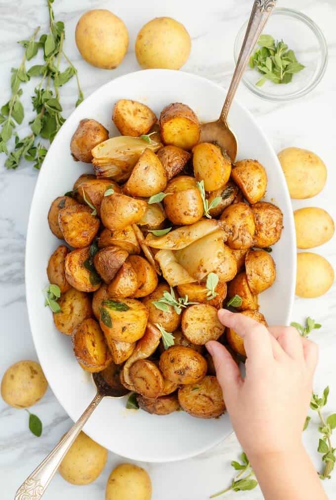 Easy Slow Cooker Breakfast Potatoes on white plate touched by hand with spoons next to herbs, scattered potatoes and bowl with more herbs
