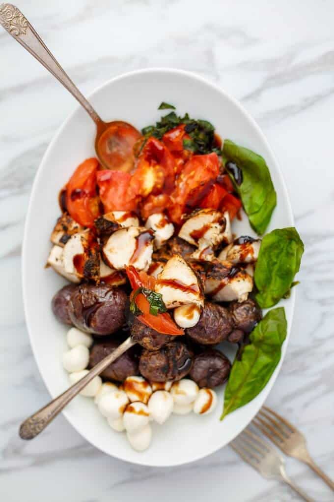Grilled Turkey Caprese Salad in white bowl with tomatoes, potatoes, mushrooms, spoons and forks on the table
