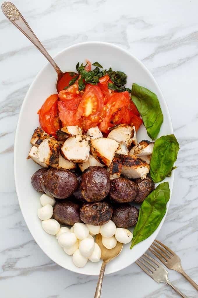 Grilled Turkey Caprese Salad in white bowl with tomatoes, potatoes, mushrooms with spoons and forks on the table