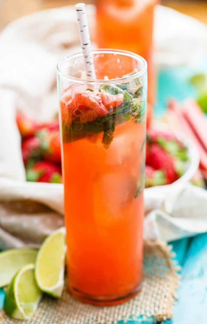 Strawberry-Rhubarb Mojitos in glass cup with straw, fresh limes and ripe strawberries in bowl on blue table