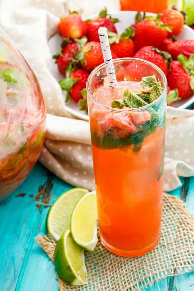 Strawberry-Rhubarb Mojitoin glass cup and pitcher with straw. Fresh limes and ripe strawberries in bowl on blue table