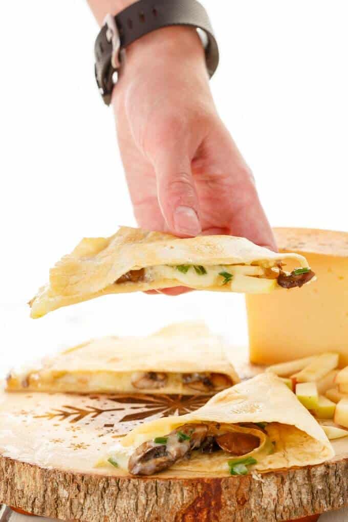 Apple-Cheese Quesadillas with Mushrooms on wooden pad and one piece held by hand. Cheese in the background