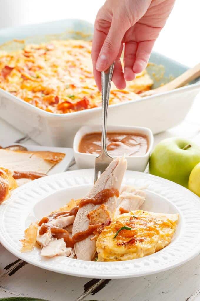 Roasted Apple Butter Turkey with Turkey Bacon Scalloped Potatoes on white plate picked by for  held by han. Casserole with same dish, bowls of sauce and apples on white table