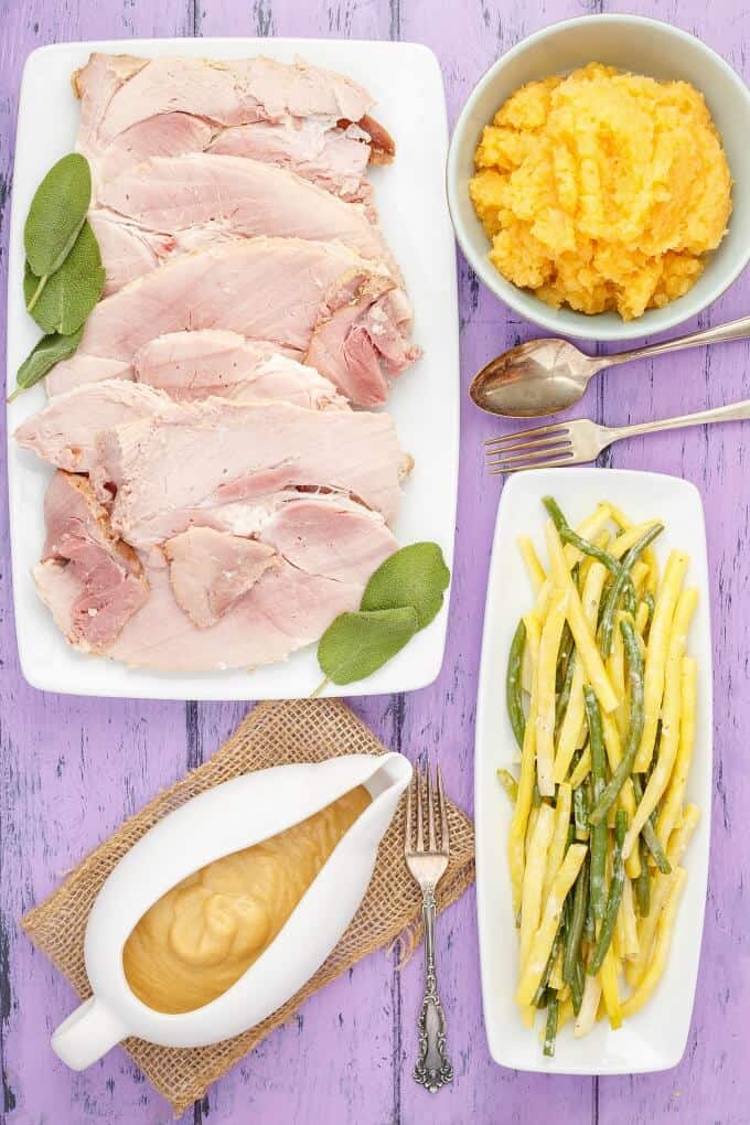 Slow Cooker Honey-Mustard Apple Ham on white tray next to bowl of side dish, tray full of vegetables, bowl of sauce,, forks and spoon on purple table