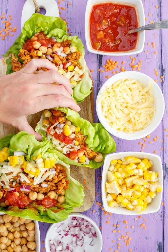 Taco Turkey-Lentil Lettuce Wraps on wooden pad touched by hand with bowls of ingredients on purple table