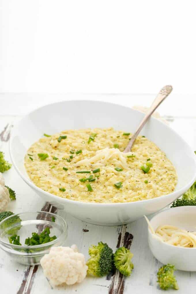 Cheesy Broccoli-Cauliflower Risotto in white bowl with spoon. Bowls with ingredients and scattered cauliflower and broccoli on white table