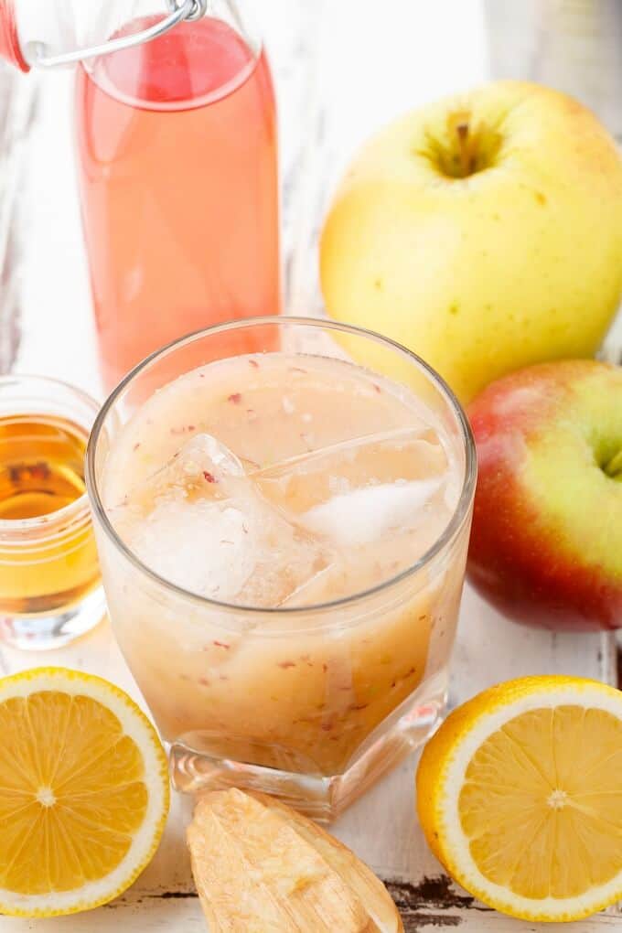 Fresh Apple Whiskey Sour in glass cup with ice. Apples, lemons, glass shot and glass bottle around.