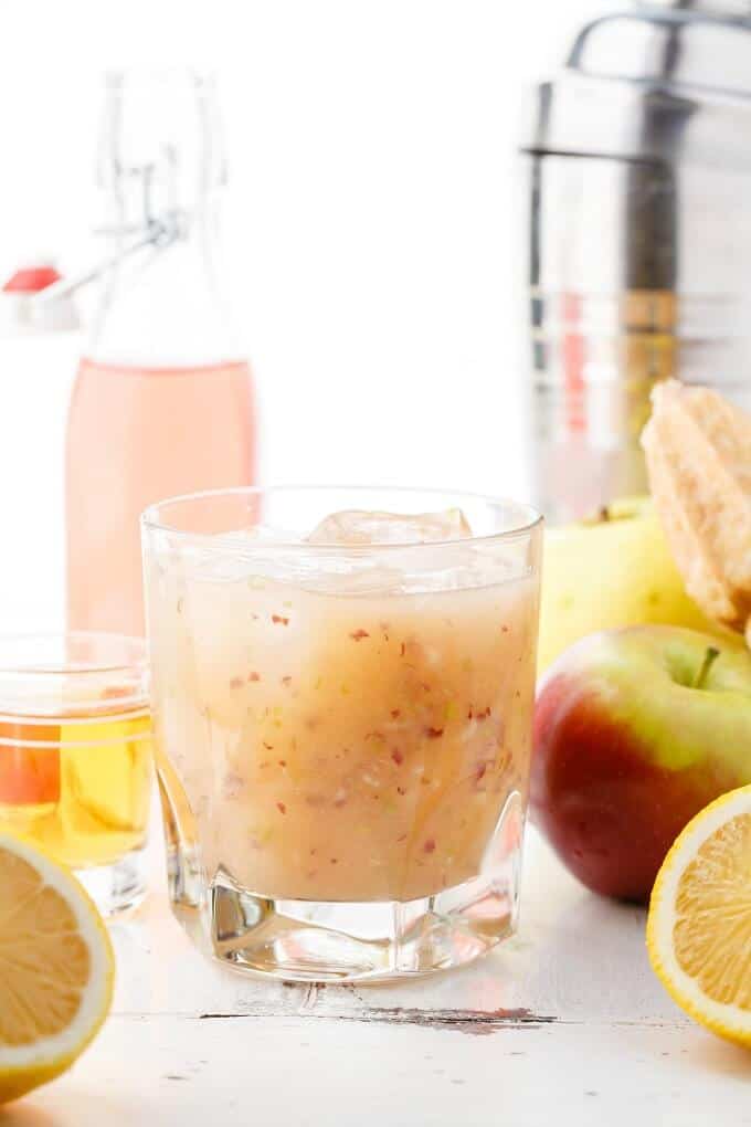 Fresh Apple Whiskey Sour in glass cup with ice. Apples, lemons, glass shot and glass bottle around.