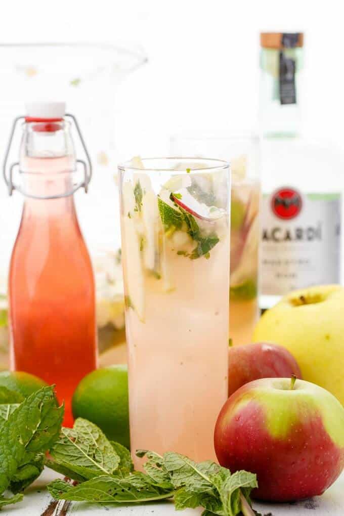 Apple Mojitos in glass cup with apples, limes, herbs, syrup glass bottle and bacardi liquor around