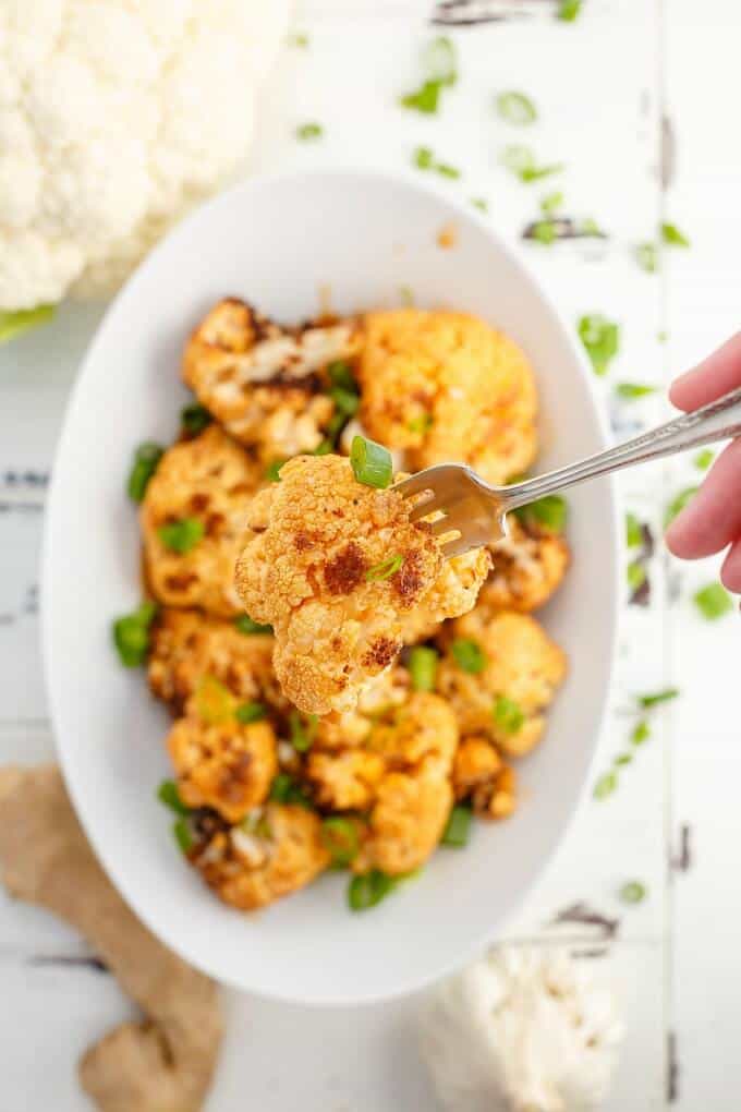 General Tso's Cauliflower picked by fork over bowl of same dish next to fresh cauliflower