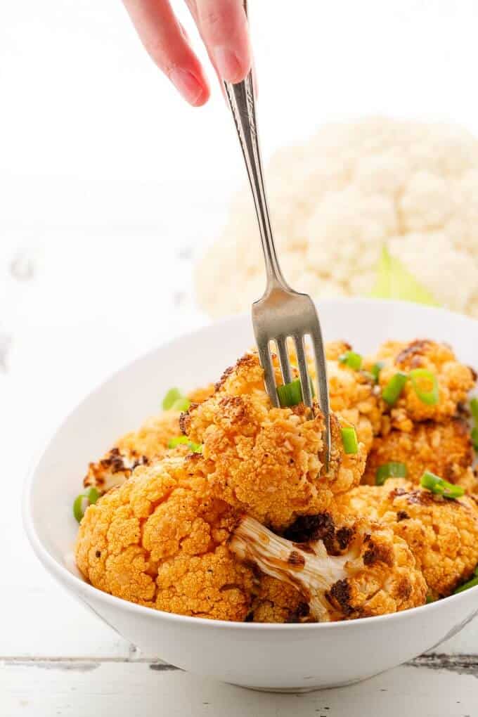General Tso's Cauliflower in white bowl with fork held by hand, cauliflower in the background