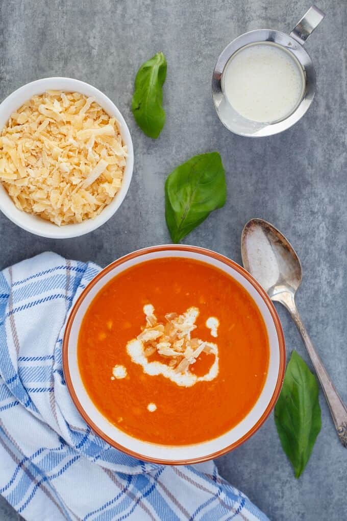 Roasted Red Pepper Soup with Gouda in orange white bowl. Spoon, herbs, jar of milk, bowl of cheese and cloth wipe on gray table