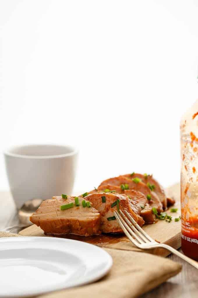 Slow Cooker Sriracha Pork Tenderloin slices on wooden pad with fork, plastic bottle, white bowl, plate and spoon