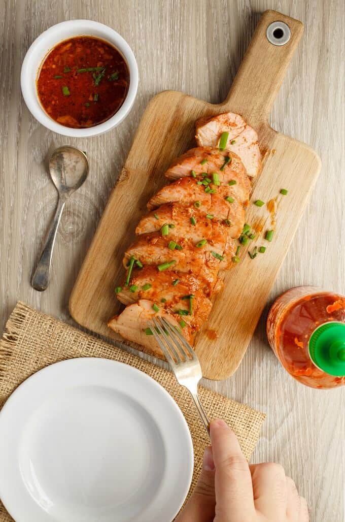 Slow Cooker Sriracha Pork Tenderloin slices on wooden pad next to plastic bottle, bowl of sauce, spoon, fork held by hand and white plate on brown table