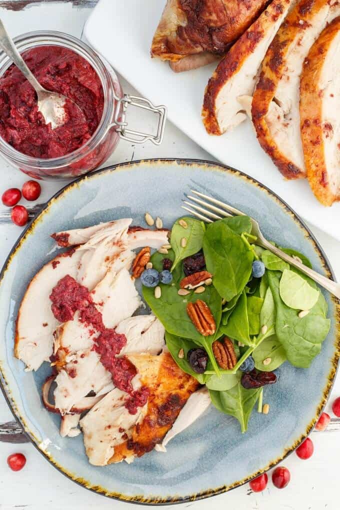 Roasted Cranberry Butter Turkey on blue plate with walnuts, herbs, blueberries, fork next to jar of butter, tray with turkey and scattered cranberries