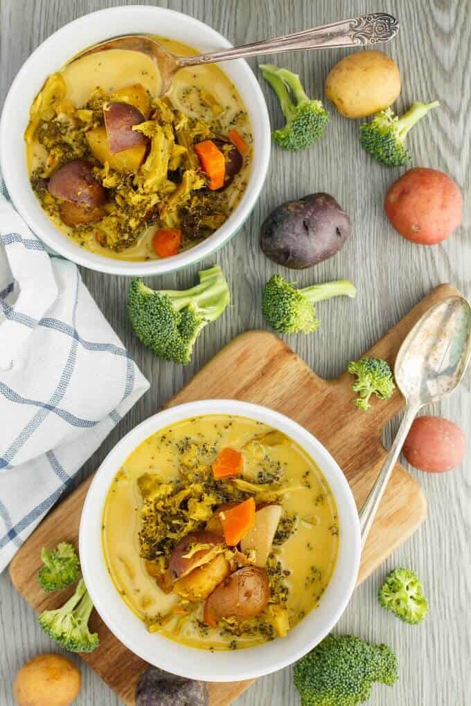 Roasted Potato-Vegetable Soup  in white bowl on wooden pad and gray table. Spoon, scattered brocolli and potatoes around table with cloth wipe