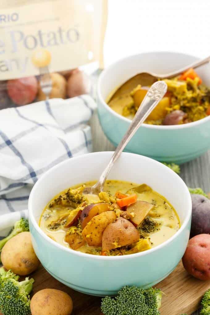 Roasted Potato-Vegetable Soup  in blue bowl with spoon on wooden pad with vegetable and potatoes around. Cloth wipe and potato sack in the background