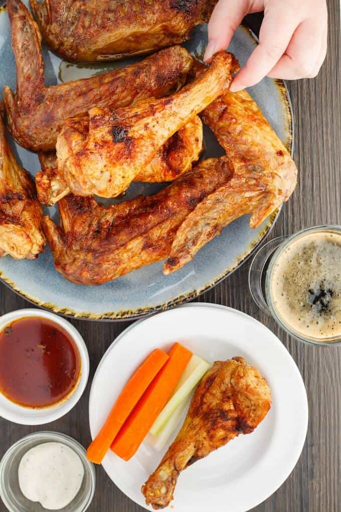 Zesty Maple Turkey Wings on blue tray touched by hand. One wing on white plate with vegetable. Sauce, ingredient in bowls and cup of coffe on gray table