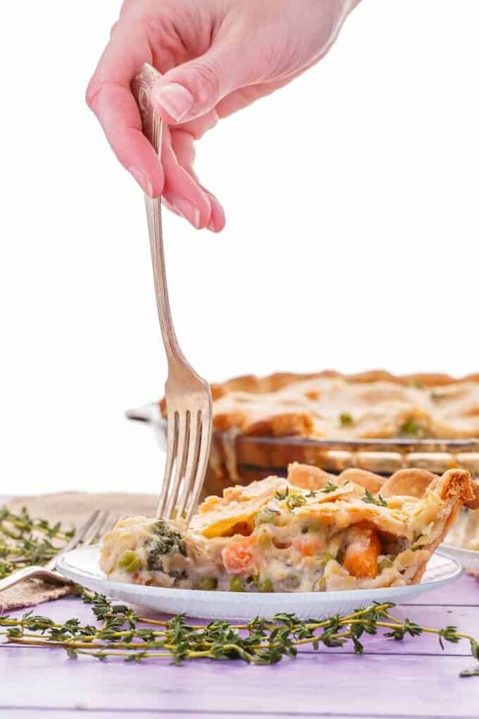 Hearty Vegetarian Pot Pie on white plate with fork held by hand with  herbs on purple table. Glass tray with pie in the background