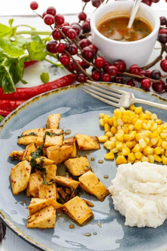 Inauthentic Thai Basil Tofu  on blue plate with fork, corn and mashed potatoes. Peppers, ,herbs, cherry tomatoes and bowl of sauce with spoon in the background