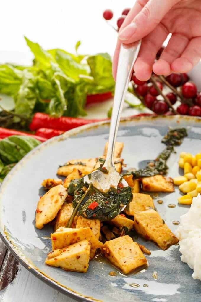 Inauthentic Thai Basil Tofu  on blue plate with spoon held by hand with corn, mashed potatoes. Vegetables and fruits around the table