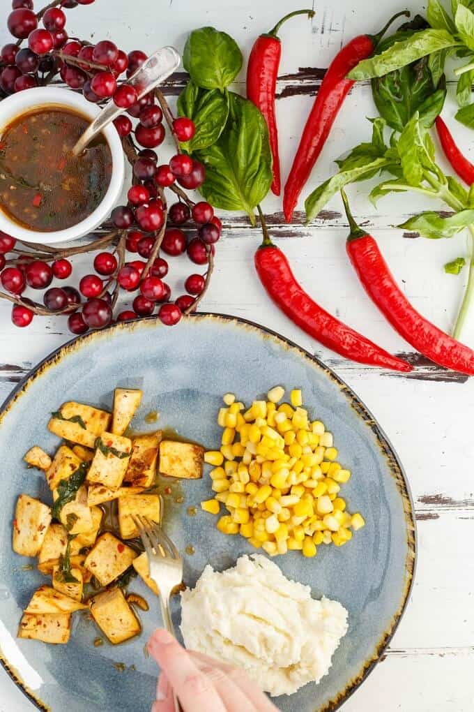 Inauthentic Thai Basil Tofu on blue plate with mashed potatoes, corn and fork held by hand. Peppers, herbs, cherry tomatoes and bowl of sauce with spoon on the table