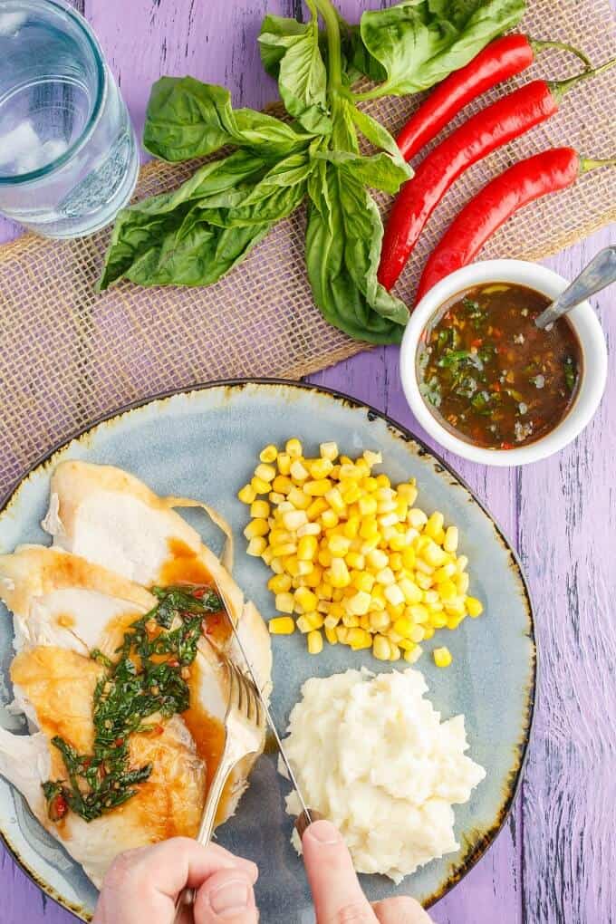 Inauthentic Thai Basil Turkey on blue plate with fork and knife held by hands with corn and mashed potatoes. Peppers, herbs, bowl of sauce and glass cup around the table