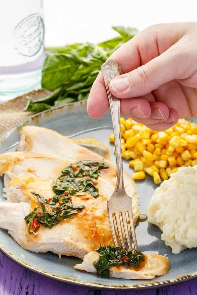 Inauthentic Thai Basil Turkey on blue plate with fork held by hand with corn, mashed potatoes, glass cup and herbs in the background