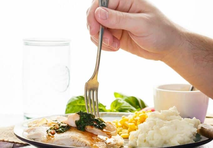 Inauthentic Thai Basil Turkey on plate with fork held by hand and mashed potatoes, corn. Glass cup, bowl with spoon and herbs in the background