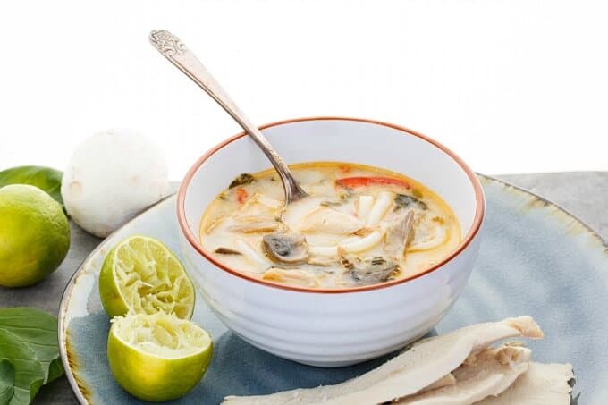 Inauthentic Thai Basil Turkey Soup  in white orange bowl with spoon on blue tray with two hlafs of lemon and leftover turkey. Lemon, mushroom and herbs on gray table