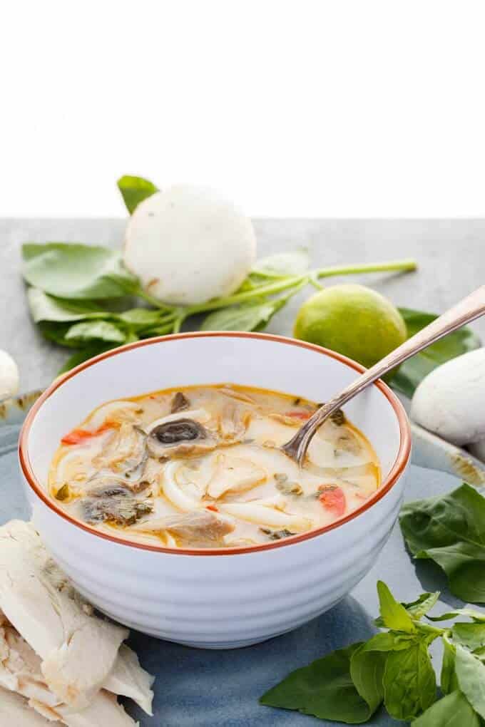 Inauthentic Thai Basil Turkey Soup  in orange white bowl witj spoon. Leftover turkey, herbs, mushrooms and lemon on gray table