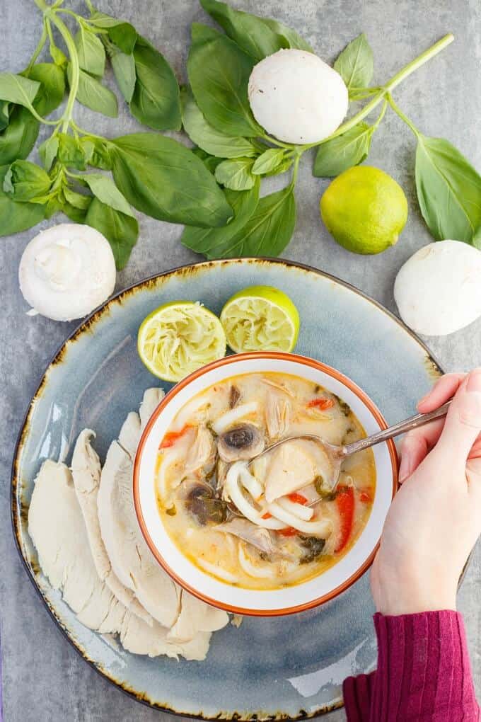 Inauthentic Thai Basil Turkey Soup  in orange white bowl with spoon held by hand on blue tray with turkey and lemon sliced in half. Mushrooms, lemon and herbs on gray table