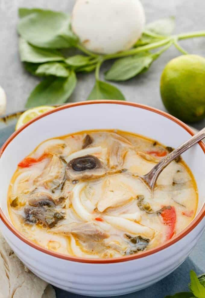 Inauthentic Thai Basil Turkey Soup  in white orange bowl with spoon. Lemon, herb mushroom on gray table