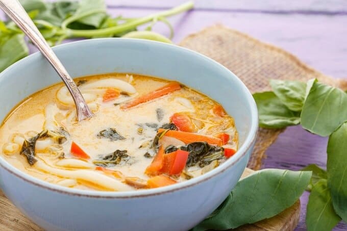 Inauthentic Vegetarian Thai Basil Soup in white bowl with spoon, herbs on purple table