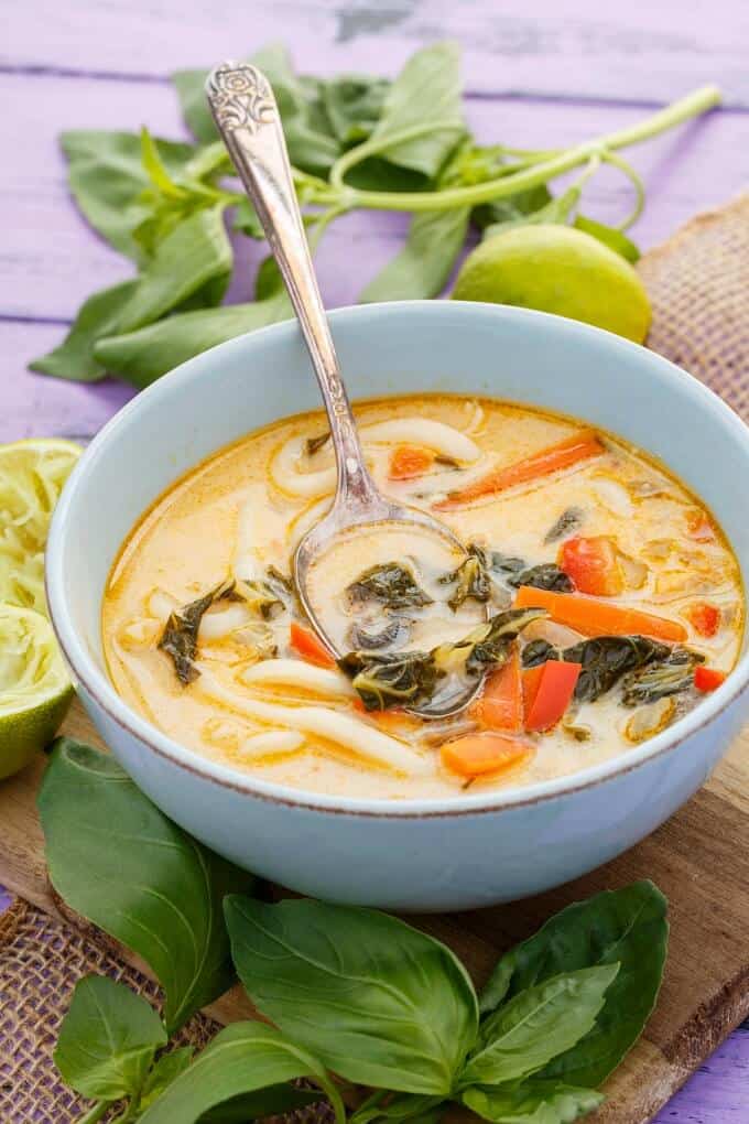 Inauthentic Vegetarian Thai Basil Soup in white bowl with spoon, herbs, lemons  on purple table