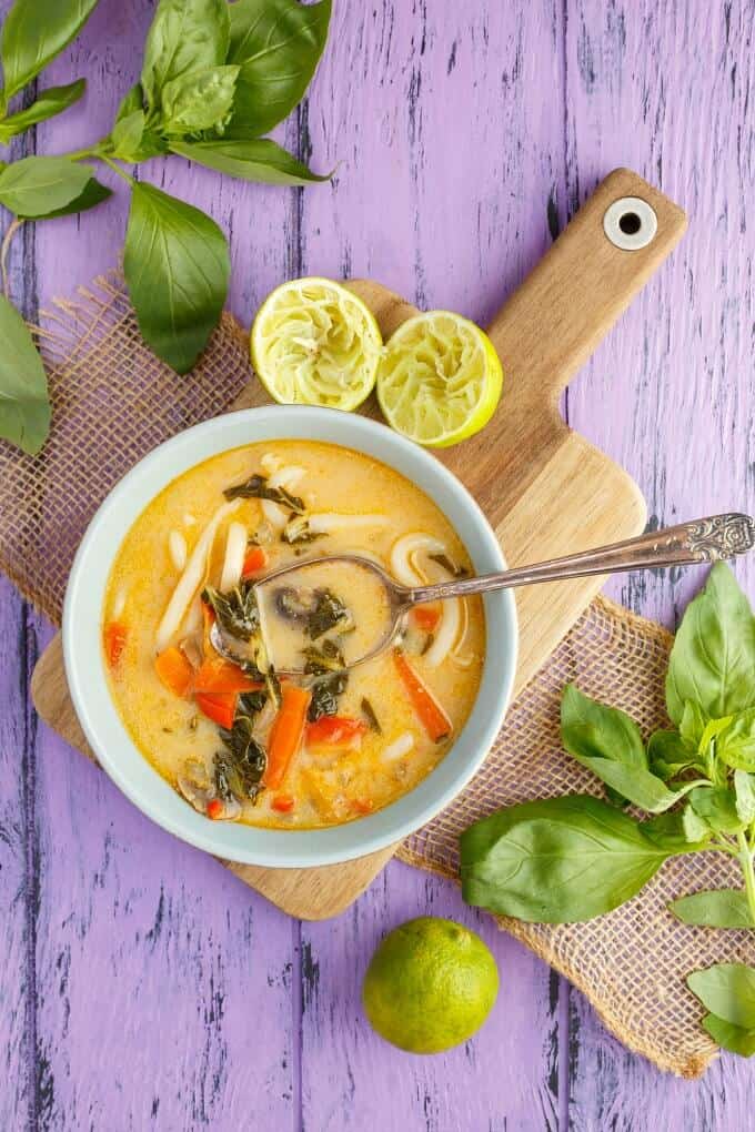 Inauthentic Vegetarian Thai Basil Soup in white bowl with spoon on wooden pad with lemon sliced in half. Herbs, lemon on purple table