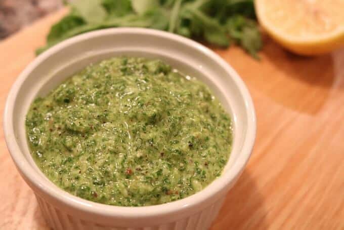 Lemony arugula pesto in pinkish bowl with herbs and lemon in the background
