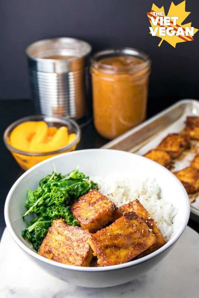 Oeach bbq baked tofu in white bowl with vegetable and rice, bowl of peaches, metal and glass jar in the background