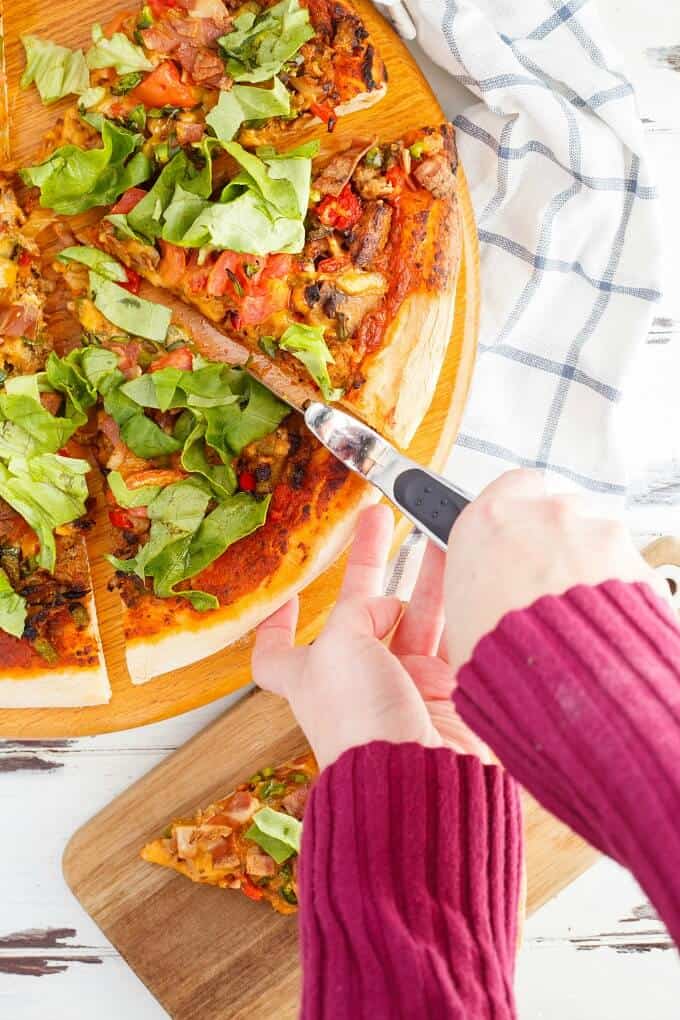 Turkey Bacon Cheeseburger Pizza on wooden pad being sliced by piza cutter held by hand