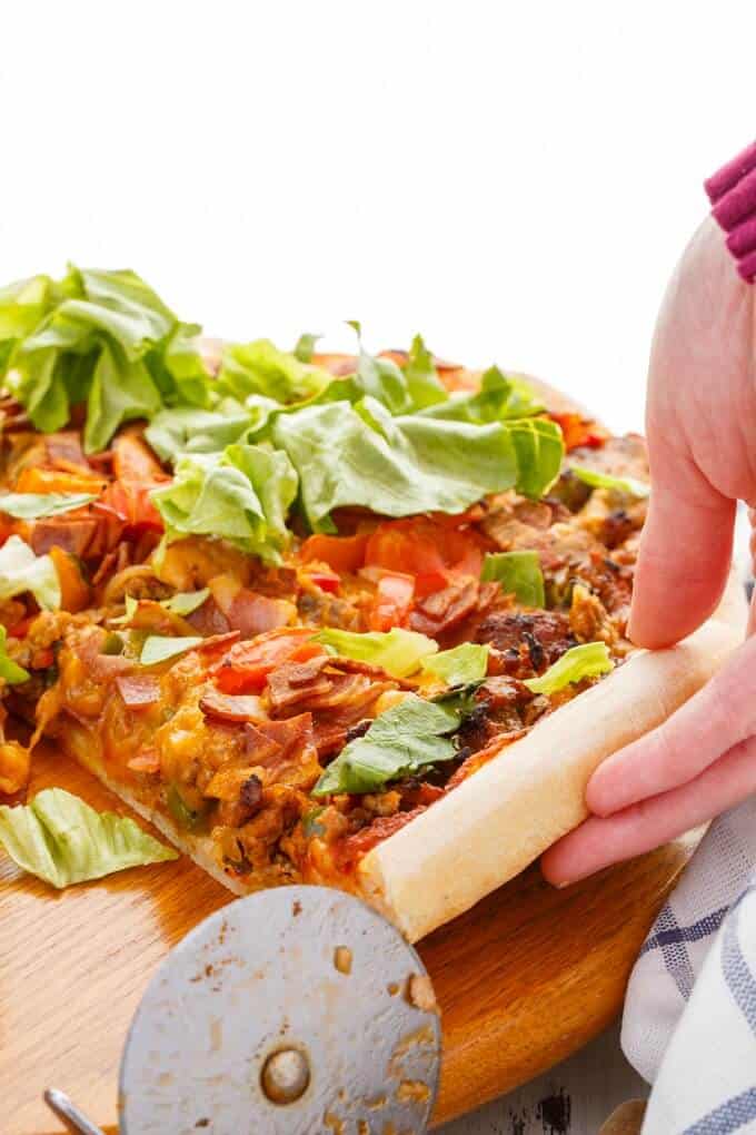 Turkey Bacon Cheeseburger Pizza on wooden pat with pizza cutter, pizza slice held by hand