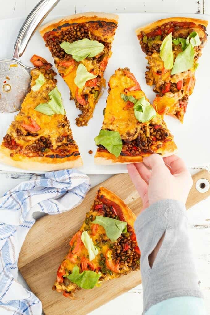 Vegetarian Lentil Cheeseburger Pizza slices on white tray with pizza cutter and on wooden pad next to cloth wipe, one slice held by hand