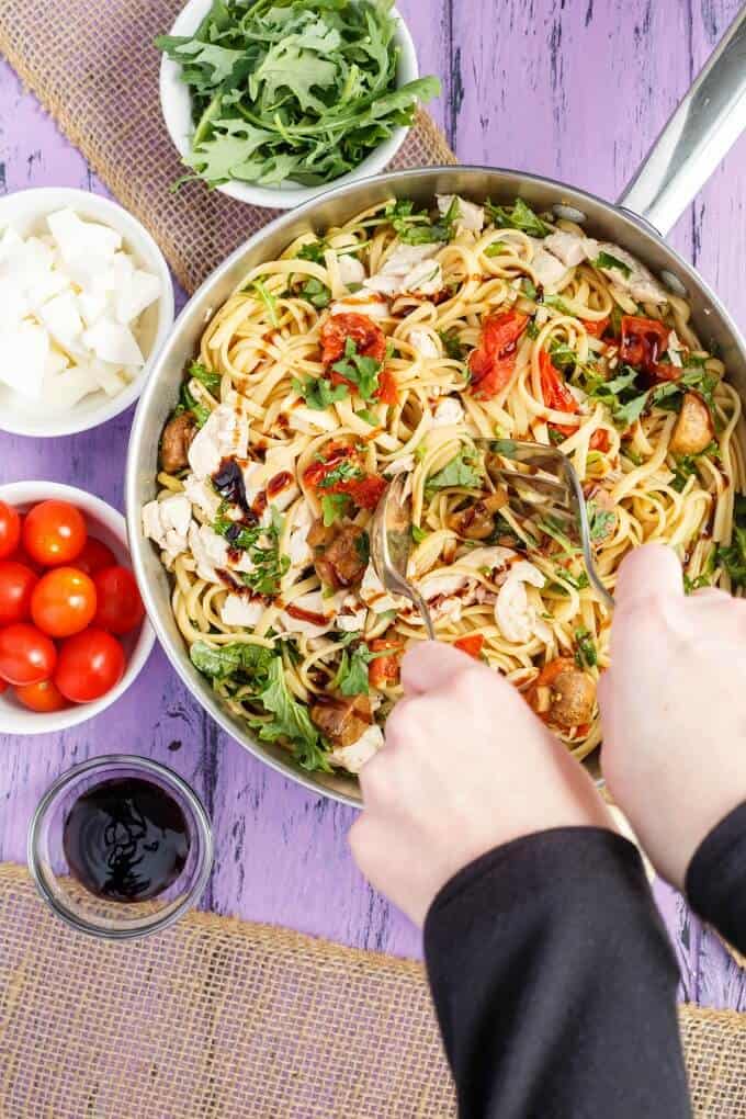 Roasted Chicken Caprese Pasta in pan mixed by forks held by hands. Bowls of tomatoes, sauce, cheese and herbs on purple table