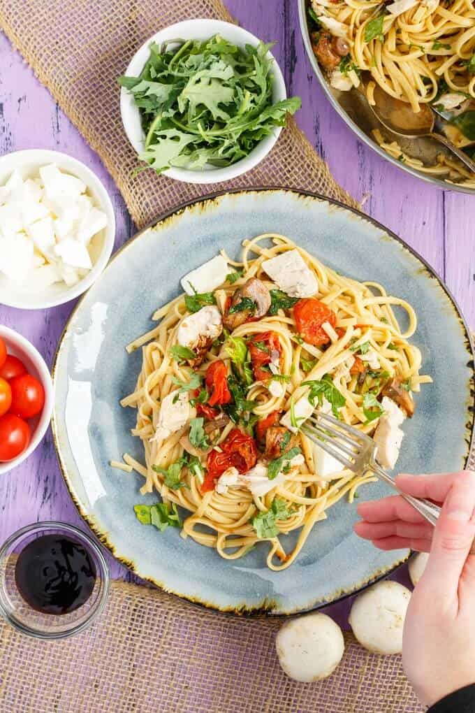 Roasted Chicken Caprese Pasta on blue plate with fork held by hand. Mushrooms, bowls with tomatoes, cheese, sauce and herbs on purple table
