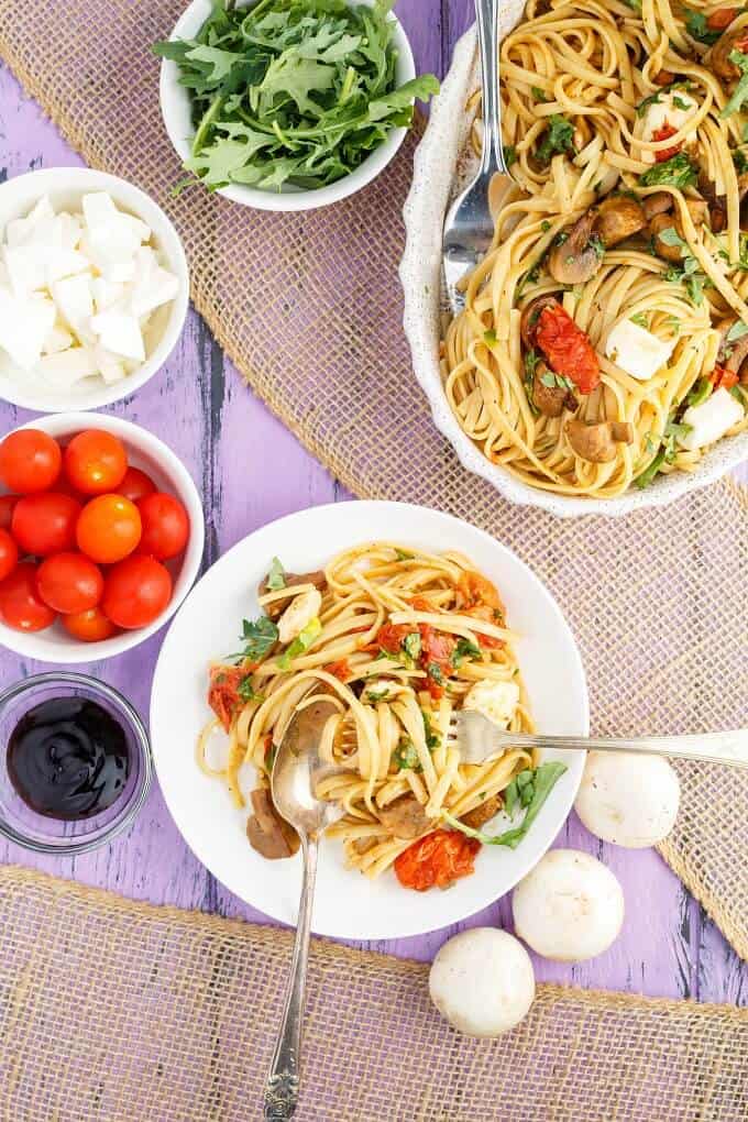 Roasted Mushroom Caprese Pasta in bow and on white palte with spoon and fork. Bowls of sauce, tomatoes, cheese, herbs on purple table with mushrooms